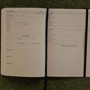 May include: An open golf logbook with white pages featuring black printed text and grids for tracking scores, course details, weather, and notes. The notebook includes a black elastic band closure that runs vertically across the right page.