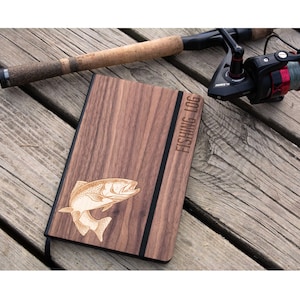 May include: A rectangular notebook with a dark brown wood grain cover featuring an engraved fish illustration and the text FISHING LOG. A black elastic band secures the cover, and a fishing rod with a cork handle and reel lies nearby.