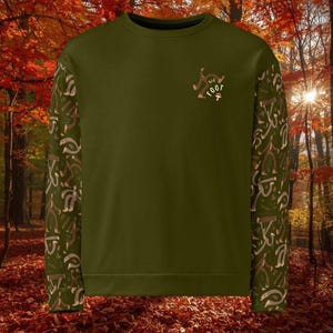 Könnte beinhalten: Olivgrünes Sweatshirt mit gemusterten Ärmeln. Die Ärmel haben ein sich wiederholendes Design in Braun- und Beigetönen. Eine kleine Grafik eines Pilzes und Text befinden sich auf der Brust. Das Sweatshirt wird vor einem Hintergrund aus Herbstlaub gezeigt.