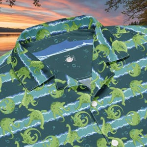 Könnte beinhalten: Knopfleiste-Hemd mit einem sich wiederholenden Muster aus grünen Seeungeheuern auf einem dunkelblauen Hintergrund mit weißen und blauen Streifen. Das Hemd hat einen Kragen und weiße Knöpfe.