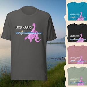 Könnte beinhalten: Ein dunkelgraues T-Shirt mit einer Cartoon-Illustration eines rosa Seeungeheuers und dem Wort "LEGENDARY". Das Seeungeheuer befindet sich im Wasser mit einem kleinen Boot. Das T-Shirt wird mit anderen Farboptionen präsentiert.