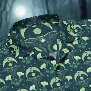 May include: Dark teal button-up shirt featuring a repeating pattern of moths, moons, and flowers in green and white. The shirt has a classic collar and button closure. The background is a blurred forest scene with a full moon.