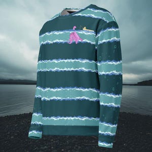 Könnte beinhalten: Ein teal- und blaues Langarm-Sweatshirt mit Wellenmuster. Das Sweatshirt zeigt eine rosa Dinosaurier-Grafik mit einer Angelrute. Der Hintergrund zeigt einen See unter einem bewölkten Himmel.