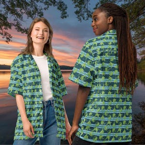 May include: Two people wearing short-sleeved button-down shirts with a repeating pattern of green dinosaurs on a dark blue background with horizontal light blue stripes. The shirts are open, revealing a white t-shirt underneath. The background shows a sunset over a lake.