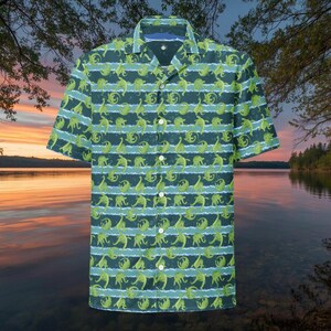 May include: Short-sleeved button-down shirt with a repeating pattern of green dinosaurs on a dark teal background. The design includes horizontal stripes of light blue. The shirt has a classic collar and white buttons.