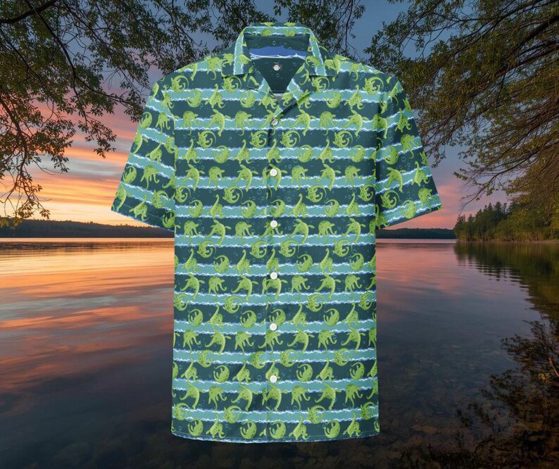 May include: Short-sleeved button-down shirt with a repeating pattern of green dinosaurs on a dark teal background. The design includes horizontal stripes of light blue. The shirt has a classic collar and white buttons.