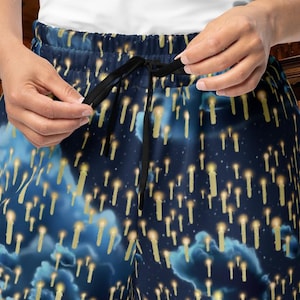 May include: Dark blue pajama pants with a repeating pattern of floating candles and stars. The pants have a black drawstring at the waist. The candles are yellow and white, and the background is a dark blue with light blue clouds.