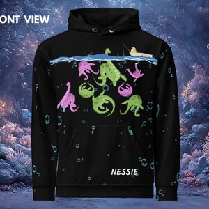 Könnte beinhalten: Schwarzer Kapuzenpullover mit Nessie-Grafik. Das Design zeigt rosa und grüne Meereskreaturen, Blasen und einen Fischer in einem Boot über der Wasserlinie. Das Wort "NESSIE" ist unten aufgedruckt.
