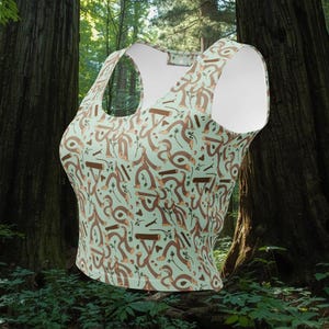 Op de afbeelding: Een mouwloze crop top met een ronde hals en een lichtgroene achtergrond. De top is bedekt met een herhalend patroon van bruine teckels, muziekinstrumenten en abstracte vormen. De top is gemaakt van een zacht, comfortabel materiaal.