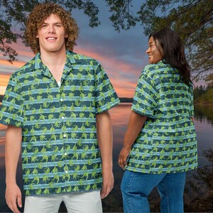 May include: Short-sleeved button-down shirt with a green dinosaur pattern on a blue and white striped background. The shirt has a classic collar and is worn by a person. The shirt is part of a matching set.