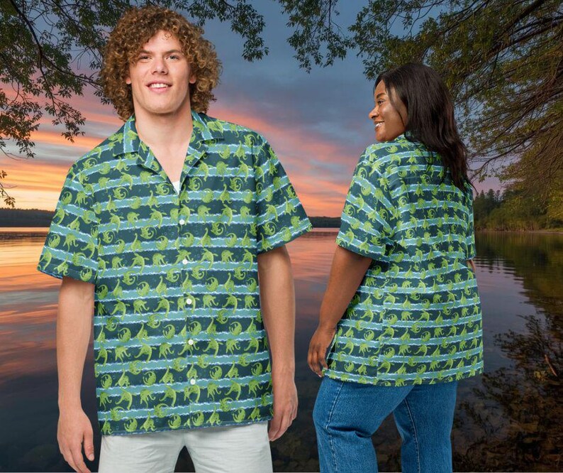 May include: Short-sleeved button-down shirt with a green dinosaur pattern on a blue and white striped background. The shirt has a classic collar and is worn by a person. The shirt is part of a matching set.