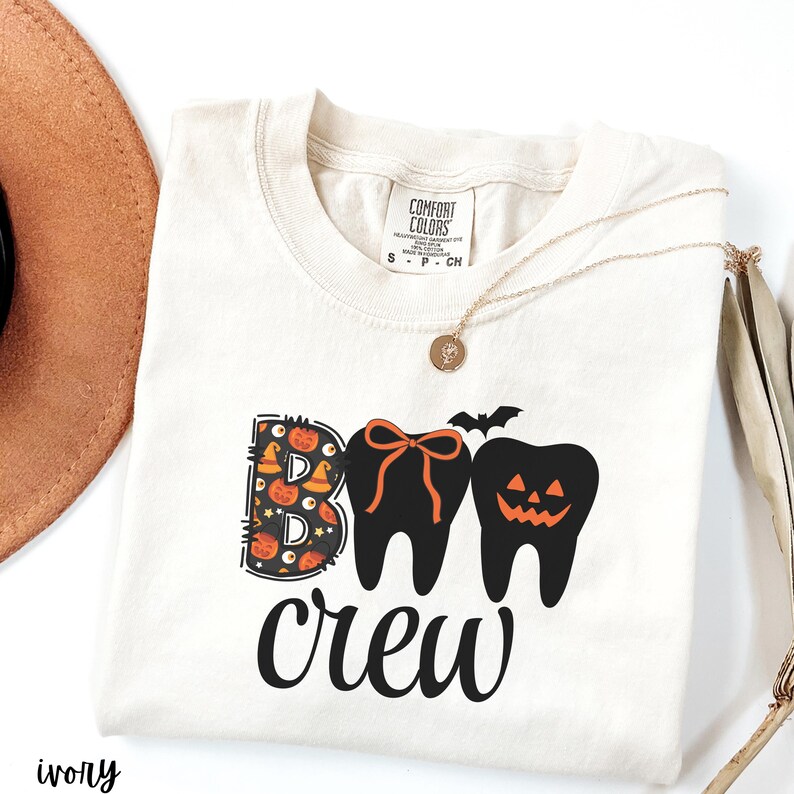 May include: Ivory-colored t-shirt with the words "BOO crew" in black. The "B" is decorated with pumpkins and bats. The "O"s are designed as teeth, one with an orange bow and the other with a jack-o'-lantern face. A gold necklace rests on the shirt.