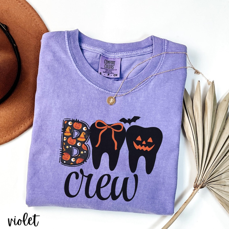 May include: A violet Comfort Colors t-shirt with a Halloween-themed graphic. The design features the word "BOO" with a pumpkin and witch hat pattern, two tooth-shaped figures, and the word "crew" in black. A gold necklace and a brown hat are also visible.