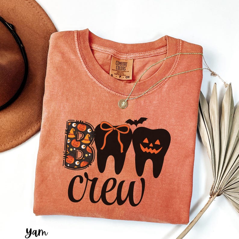 May include: A coral-colored t-shirt with the words "Boo Crew" in black script. The design features two teeth, one with a bow and the other with a jack-o'-lantern face, and a bat. The letter "B" is decorated with Halloween elements.