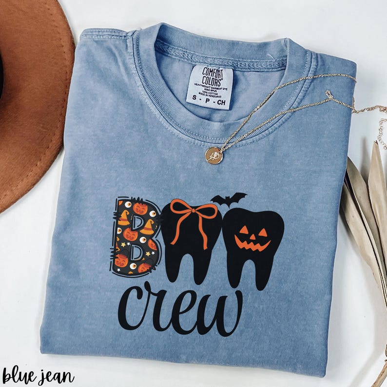 May include: A blue jean Comfort Colors t-shirt with a Halloween-themed graphic. The graphic features the word "BOO" with a pumpkin and witch hat design, and two tooth designs with a bow and jack-o'-lantern face. The word "crew" is written below.