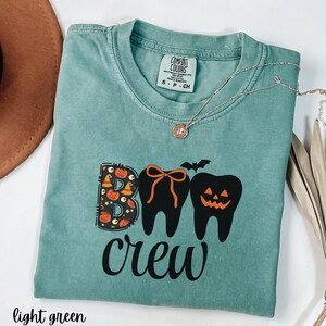May include: Light green t-shirt with the words "BOO crew" in black. The "BOO" is designed with Halloween-themed elements, and the "O"s are represented by teeth. A silver necklace is draped across the shirt.