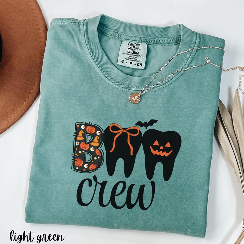 May include: Light green t-shirt with the words "BOO crew" in black. The "BOO" is designed with Halloween-themed elements, and the "O"s are represented by teeth. A silver necklace is draped across the shirt.