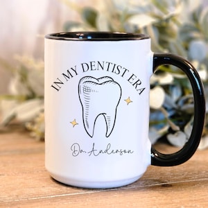 May include: White ceramic coffee mug with a black rim and handle. The mug has a black and white illustration of a tooth with the text "In My Dentist Era" and "Dr. Anderson".
