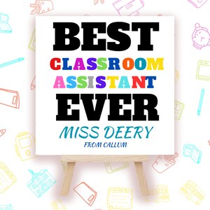 May include: A white canvas sign with a wooden easel reads "BEST CLASSROOM ASSISTANT EVER MISS DEERY FROM CALLUM". The words "CLASSROOM ASSISTANT" are in rainbow colours.