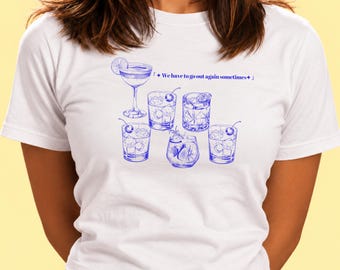 May include: A white short-sleeve t-shirt featuring a blue graphic print of various cocktail glasses filled with ice and garnishes, accompanied by the text "We have to go out again sometimes" with small star symbols on either side.