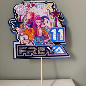 May include: A vibrant cake topper with three anime-style figures, the number 11, and the name FREYA. The topper has a blue and silver border with glitter accents and the word "HUNTRIX" at the top. A wooden skewer is attached to the bottom.
