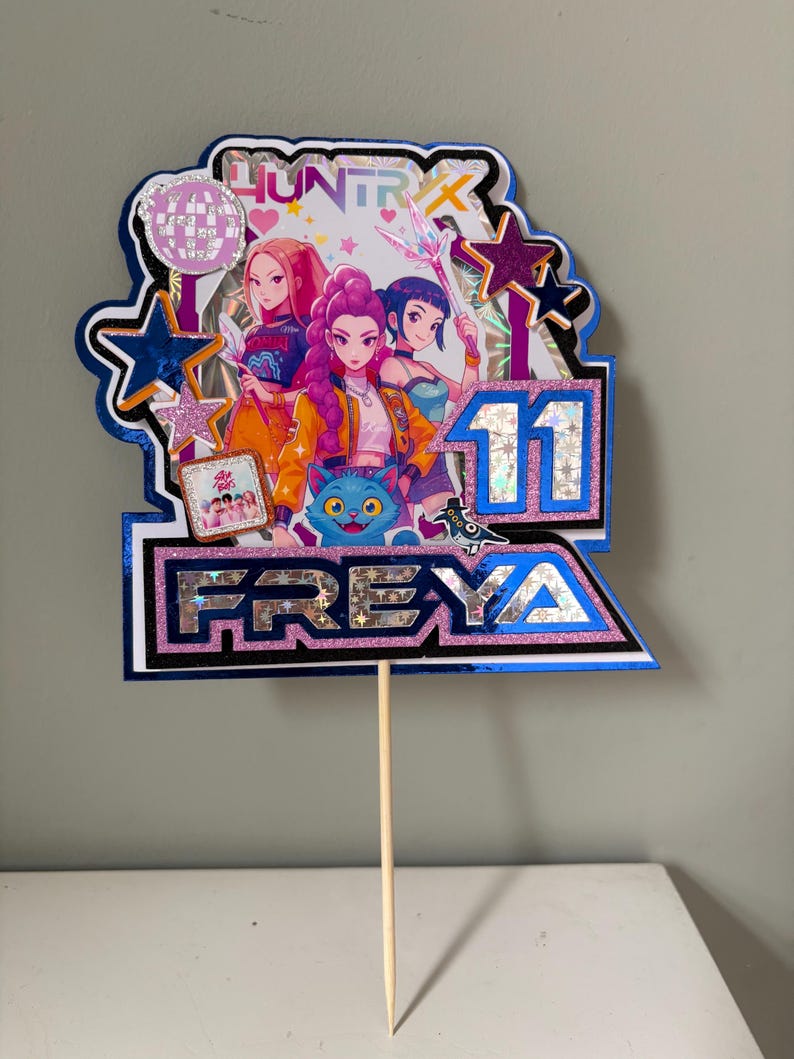 May include: A vibrant cake topper with three anime-style figures, the number 11, and the name FREYA. The topper has a blue and silver border with glitter accents and the word "HUNTRIX" at the top. A wooden skewer is attached to the bottom.
