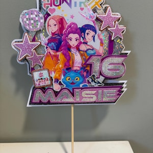 May include: A vibrant cake topper with anime-style illustrations of three figures, stars, and the text "HUNTR" and "MAISIE 16". The design incorporates a disco ball, a blue cat, and a small framed photo. The topper is attached to a wooden skewer.