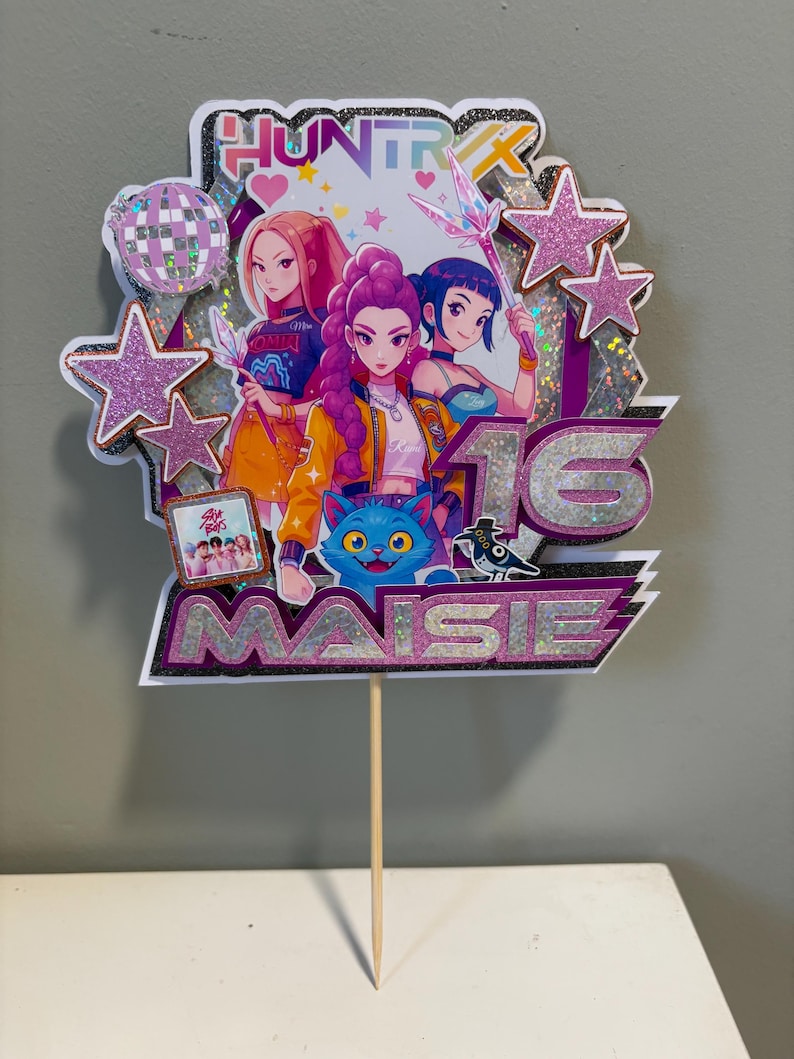 May include: A vibrant cake topper with anime-style illustrations of three figures, stars, and the text "HUNTR" and "MAISIE 16". The design incorporates a disco ball, a blue cat, and a small framed photo. The topper is attached to a wooden skewer.