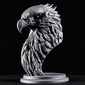 May include: A detailed gray 3D printed sculpture of an eagle's head, with a textured surface that resembles feathers. The eagle's head is turned to the side, and its beak is closed. The sculpture is mounted on a round base.