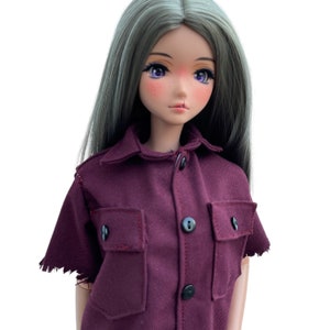 May include: A doll wearing a maroon button-up shirt with two flap pockets and frayed edges on the sleeves.