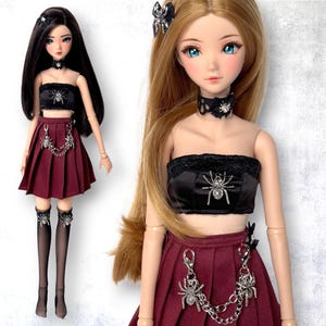 Puede incluir: Se muestran dos muñecas de moda, cada una con cabello largo y maquillaje detallado. Llevan conjuntos a juego: tops de tubo negros con adornos de arañas, faldas plisadas burdeos y accesorios con temática de arañas. Una muñeca tiene cabello negro, la otra rubio.