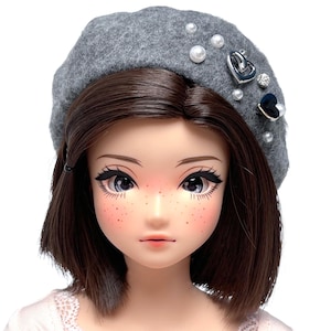 May include: A doll with brown hair and freckles wears a gray beret adorned with pearls and heart-shaped charms. The doll has large eyes with long eyelashes and is wearing a light pink top. The beret is decorated with pearls and heart-shaped charms.