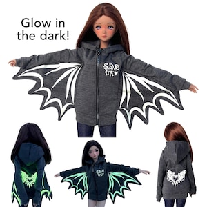 May include: A dark gray zip-up hoodie with bat wing designs that glow in the dark. The hoodie features white wing accents, a small logo on the chest, and the text "Glow in the dark!" at the top. The image shows the hoodie on a doll.