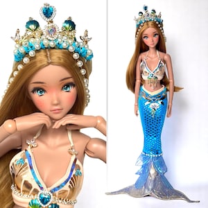 May include: A mermaid doll with long blonde hair, wearing a blue and gold seashell-themed top and a blue scale-patterned mermaid tail. The doll has a jeweled crown and a matching seashell-themed top. The doll is approximately 12 inches tall.