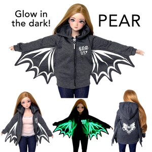 May include: A doll wearing a dark gray zip-up hoodie with white bat wing designs on the sleeves. The hoodie has a small logo on the chest and the text "PEAR" is visible. The words "Glow in the dark!" are also present.