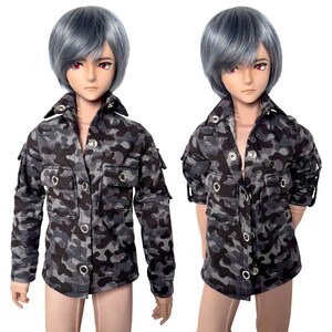 Könnte beinhalten: Zwei Puppen tragen eine grau-schwarze Tarnjacke. Die Jacke hat mehrere Taschen und silberne Knopfverschlüsse. Die Puppen haben graues Haar und rote Augen. Die Jacke ist im militärischen Stil gehalten.