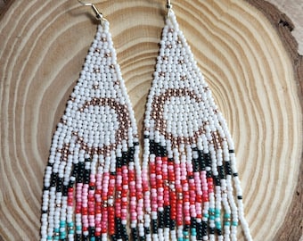 Puede incluir: Pendientes de cuentas hechos a mano con forma triangular. Los pendientes presentan un fondo blanco con un diseño floral en tonos rosa, rojo, negro y turquesa. Los pendientes se exhiben sobre una superficie de madera.