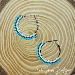 May include: A pair of silver hoop earrings with a beaded design. The beads are arranged in a pattern of turquoise, white, and green. The earrings are displayed on a wooden surface.