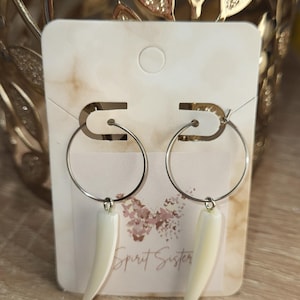 May include: Silver hoop earrings with a white, tooth-shaped pendant. The earrings are displayed on a white card with the text "Spirit Sister" and "Spirit Sisters Jewelry". The background is a blurred, golden color.