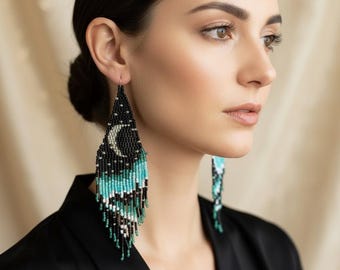 Könnte beinhalten: Lange, dreieckige Perlenohrringe mit schwarzem Hintergrund, einem weißen Halbmond und Sternenmuster oben, das in ein türkisfarbenes und schwarzes Fransendesign unten übergeht. Der Text Spirit Sisters Jewelry ist sichtbar.