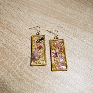Puede incluir: Un par de pendientes rectangulares dorados. Cada pendiente presenta un rectángulo relleno de resina transparente con copos de oro y detalles en rosa y rojo. Los pendientes tienen cierres de gancho dorados.
