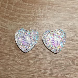 May include: Two heart-shaped embellishments with iridescent rhinestones. Each heart features a central design with a turtle motif in shades of purple and white. The rhinestones sparkle, creating a dazzling effect. The background is a wooden surface.