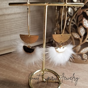 May include: A pair of gold hoop earrings with white feather accents. The earrings are displayed on a gold stand. The text "Inuit Spirit Jewelry" is visible below the stand.