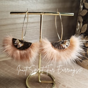 May include: A pair of gold hoop earrings with a light brown fur pom-pom and gold accents. The earrings are displayed on a gold stand. "Inuit Spirit Fur Earrings" is written below the stand.
