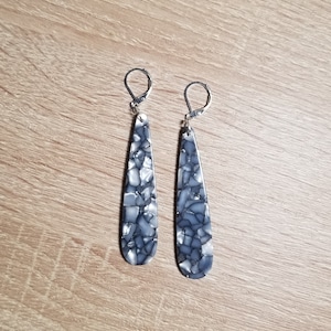 Puede incluir: Un par de pendientes largos en forma de lágrima con un gancho de metal plateado. Los pendientes están hechos de un material blanco y gris moteado.