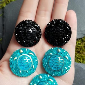 Puede incluir: Dos botones redondos negros y tres botones redondos de color turquesa, cada uno con un patrón central de caparazón de tortuga rodeado por un borde texturizado. Los botones tienen un acabado brillante y una forma circular con relieve.