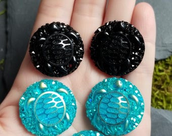 Puede incluir: Dos botones redondos negros y tres botones redondos de color turquesa, cada uno con un patrón central de caparazón de tortuga rodeado por un borde texturizado. Los botones tienen un acabado brillante y una forma circular con relieve.