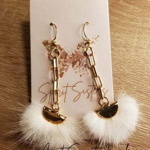 May include: A pair of gold chain earrings with white fluffy pom-poms and gold half-circle accents. The earrings are displayed on a white card with the text "Spirit Sisters" and "Inuit Spirit Jewelry".