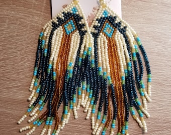Puede incluir: Un par de pendientes largos de cuentas con un patrón geométrico en tonos de azul, marrón, blanco y turquesa. Los pendientes tienen un diseño de flecos y cuelgan de ganchos dorados.