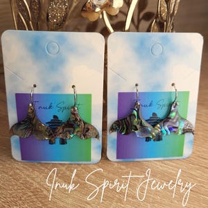 May include: Two pairs of abalone shell earrings with silver hooks, displayed on cards with a blue, purple, and green gradient. The earrings have a fan-like shape with iridescent colors. The text "Inuk Spirit Jewelry" is written in cursive.